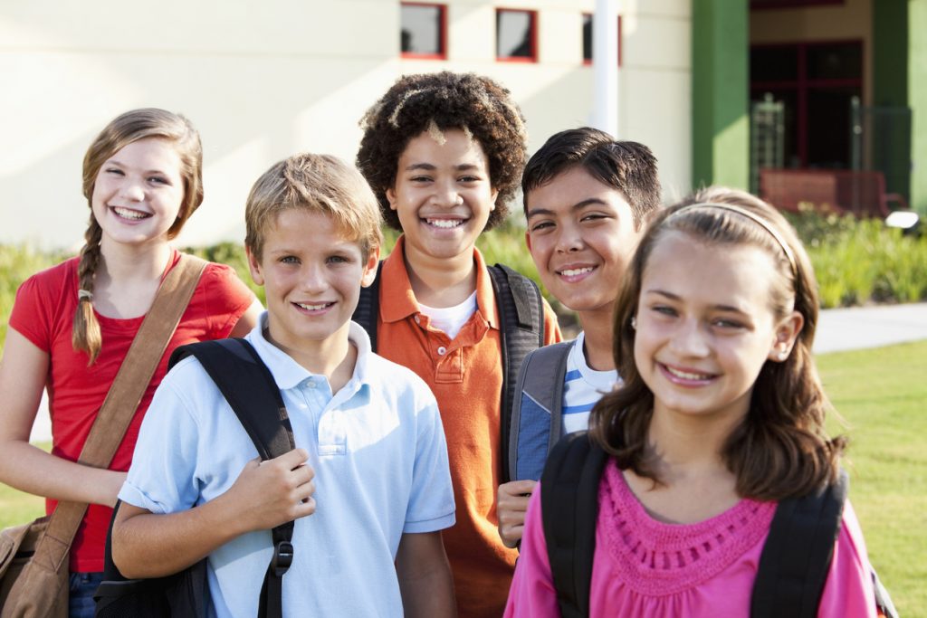 A multi-ethnic group of five elementary or middle school students standing outside a school building.  The focus is on the boys in the middle row.  The children are 10 to 12 years old.  They are smiling at the camera, all carrying bookbags or backpacks.  It is a bright, sunny day.